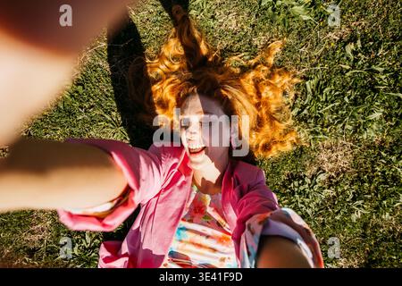 Lächelnde Frau mit rotem Haar, die auf grünem Gras liegt und an einem sonnigen Tag ein Selfie macht Stockfoto