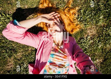 Rothaarige Frau, die auf grünem Gras liegt, lächelt und ihre Augen vor der hellen Sonne schützt Stockfoto