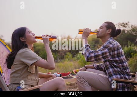 Glückliches asiatisches Paar, das bei einem Sommercampingausflug zur goldenen Stunde mit Bierflaschen toast Stockfoto