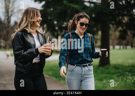 Zwei Frauen laufen in einem Park, lächeln und halten Kaffeetassen, während sie sich unterhalten Stockfoto