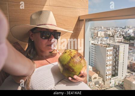 Frau, die sich auf einem Balkon entspannt und Kokoswasser mit Blick auf die Stadt trinkt Stockfoto