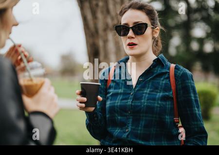 Frau in kariertem Hemd mit Kaffee draußen, Sonnenbrille und Umhängetasche, die mit einem Freund spricht Stockfoto