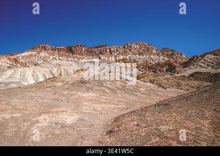 Zerklüftete Klippen erheben sich steil gegen einen leuchtend blauen Himmel. Trockene Erdhänge führen zu alten, verwitterten Steinformationen. Sonnenlicht wirft scharfe Schatten Akro Stockfoto