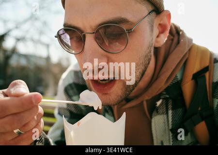 Junger Mann isst Eis aus einer Tasse zum Mitnehmen, während er bei sonnigem Wetter auf einer Parkbank sitzt. Konzept von Streetfood, Freizeit, Freizeitgestaltung und Stockfoto