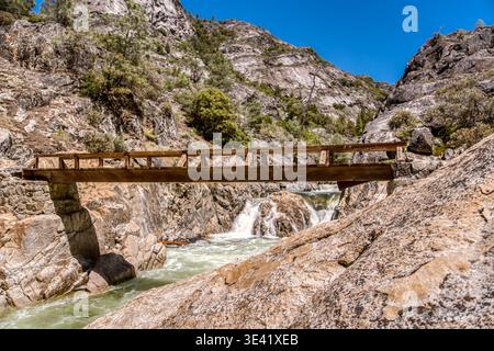 Die robuste Brücke überspannt rauschendes Wasser. Sonnenlicht leuchtet von den felsigen Wänden des Canyons. Leuchtend grüne Bäume schmiegen sich an steile Hänge. Klare blaue Himmelsunterschiede Stockfoto