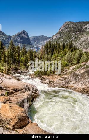 Der rauschende Fluss fließt über glatte Felsen. Waldbäume säumen den Wasserrand. Berge erheben sich hinter dem Grün. Klarer blauer Himmel umrahmt die Szene. Natur Stockfoto