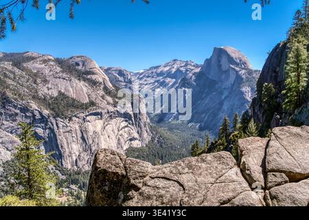 Massive Granitgipfel erheben sich unter dem klaren blauen Himmel. Schwache Pfade schlängeln sich durch Täler, eingerahmt von alten Kiefern. Schroffe Felsbrocken sitzen im Vordergrund, Addi Stockfoto