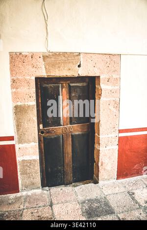 Verwitterte Holztür in rauem Steinrahmen. Rote und cremefarbene Wände umrahmen den alten Eingang. Der Boden aus Schachbrettstein sorgt für rustikalen Charme. Dunkle Holzpfanne Stockfoto