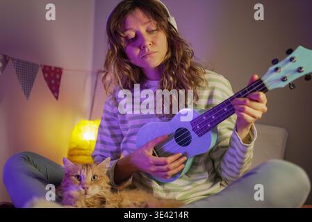 Junge Frau, die Ukulele übt, während sie Kopfhörer trägt, Musik in ihrem Schlafzimmer kreiert, zusammen mit einer einheimischen Ingwerkatze, die beobachtet, Freude und Verbindung durch ihr Musikinstrument findet Stockfoto