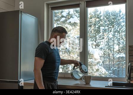 Senior man gießt Wasser aus dem Wasserkocher in einen Becher und macht seinen Morgenkaffee oder Tee in der Nähe des Winterfensters. Kalter Wintermorgen, tägliche Routine, Alleinleben Konzept Stockfoto