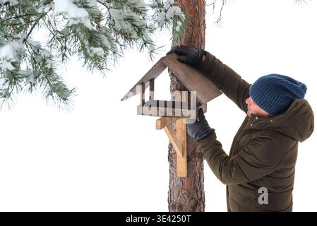 Mann mittleren Alters macht hölzerne Vogelfuttermittel auf Kiefer im Winter, weißer Schnee Hintergrund, Kopierraum für Text. Fütterung von Vögeln und Eichhörnchen in kalter Winte Stockfoto