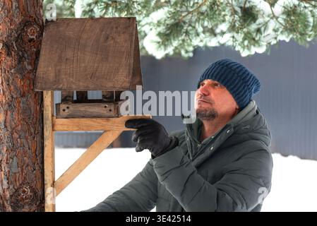 Ein Mann mittleren Alters macht und hängt im Winterpark hölzerne Vogelfutterhäuschen an Kiefern. Fütterung von Vögeln und Eichhörnchen im kalten Winter. Aktiver Abgang. Stockfoto