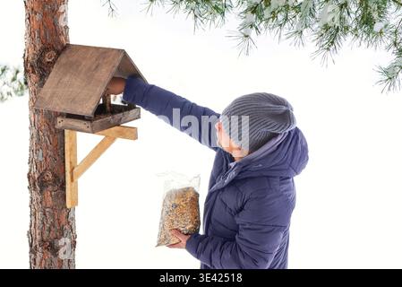 Mann mittleren Alters füllen Holzvogelfutter mit Samen auf Kiefer im Winterpark auf. Fütterung von Vögeln und Eichhörnchen im kalten Winter. Aktiver Abgang und Stockfoto