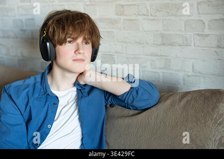 Ernsthafter junger Mann in Kopfhörern, der zu Hause etwas auf dem Sofa hört. Hören Sie sich einen Podcast, eine Vorlesung oder ein Hörbuch an Stockfoto