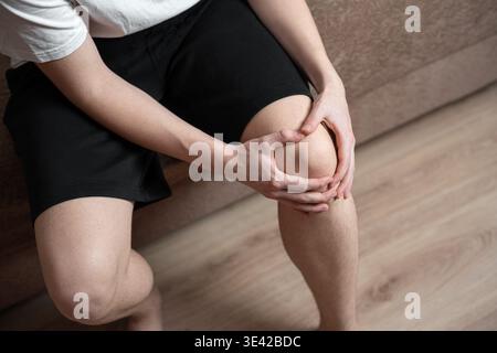 Nahaufnahme eines Mannes, der sein Knie in Unwohlsein hält. Sie leiden an Knieschmerzen, während Sie zu Hause auf dem Sofa sitzen. Gesundheitskonzept, Knieverletzung oder Arthritis c Stockfoto