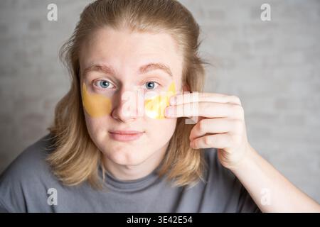 Junger gutaussehender blonder Mann, der gelbe Augenflecken aufträgt, Nahaufnahme Porträt. Beauty-Routine, mit feuchtigkeitsspendenden Gesichtspflecken. Stockfoto