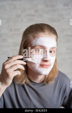 Junger blonder Mann, der kosmetische Ton Gesichtsmaske aufträgt. Grooming-Enthusiast nach täglicher männlicher Hautpflege für klare Haut. Stockfoto