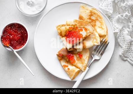 Dünne Crepes mit rotem Kaviar und Creme auf weißem Teller, Blick von oben. Herzhafte Pfannkuchen mit Lachsfischkaviar. Maslenitsa-Frühstück für traditionelles Frühstück Stockfoto