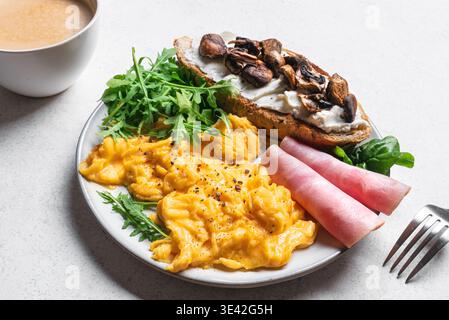 Gesundes, ausgewogenes Frühstück. Rührei, grüne Salatmischung, Schinken, Sandwich mit Frischkäse und gebratenen Pilzen. Stockfoto