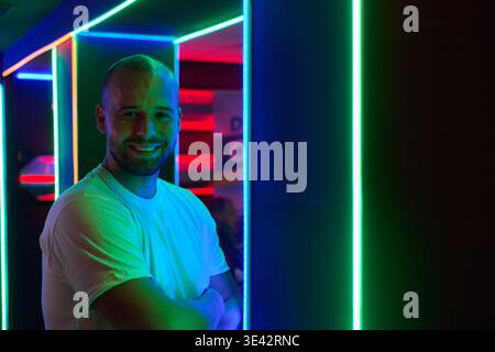 Lächelnder Mann, der im Neon-LED-Lichtkorridor im Nachtclub mit farbenfroher Umgebungsbeleuchtung steht Stockfoto