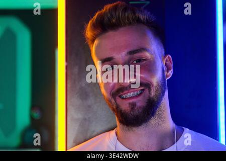 Junger Mann mit Zahnspangen lächelnd in Neon Porträt, lässigem Bart und farbenfroher LED-Beleuchtung Stockfoto