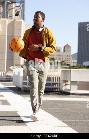 Afroamerikanischer Mann, der auf dem Dach läuft und Basketball hält, Senfjacke und Kopfhörer trägt Stockfoto