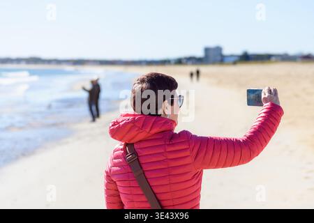 Frau in roter Jacke, die an einem sonnigen Herbsttag ein Selfie am Sandstrand macht. Unscharfer Hintergrund. Stockfoto