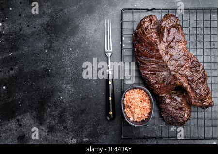 Gegrilltes Rindfleischbügel Steakonglet auf Metallgestell mit Gabel und grobem Salz, dunkler Steinhintergrund mit Kopierraum für Grillkonzept Stockfoto