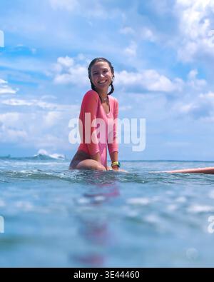 Junge Surferin, Die An Bord Sitzt Und Lächelnd Wartet, Inmitten Des Ruhigen Ozeans Und Des Blauen Himmels. Violetter Rashguard, Sanft Stockfoto