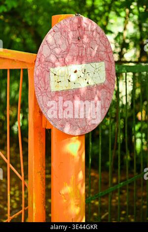 Altes, verwittertes Schild „No Entry“ mit gerissener Farbe an einem rostigen Tor. Vintage Straßenschild Hintergrund mit Sonnenlicht Schatten und grünem Parklaub Stockfoto