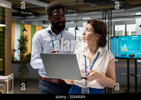 Schwarzer Mann und weiße Frau diskutieren über Geschäftsanalysen und Investitionsberichte auf Notebooks. Kollegen aus verschiedenen Ethnien konzentrieren sich auf Unternehmenslösungen, Teamarbeit und Unternehmenswachstum. Stockfoto
