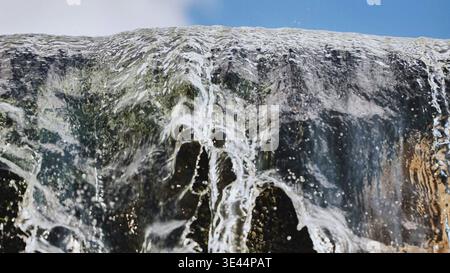 Dynamische Nahaufnahme von klarem Wasser, das über einen moosigen Rand vor einem blauen Himmel fließt. Stockfoto