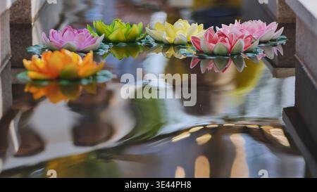 Bunte künstliche Lotusblüten, die in einer Reihe auf plätscherndem Wasser mit ruhigen Reflexen schweben Stockfoto