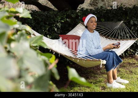 Afroamerikanerin, die auf einer Hängematte im Garten sitzt, einen Becher mit Weihnachtsmütze hält, Kopierraum Stockfoto