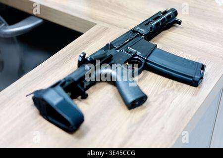 Schwarzes Gewehr im AR-15-Stil mit abnehmbarem Magazin, das auf einem Holztisch im Büro liegt Stockfoto