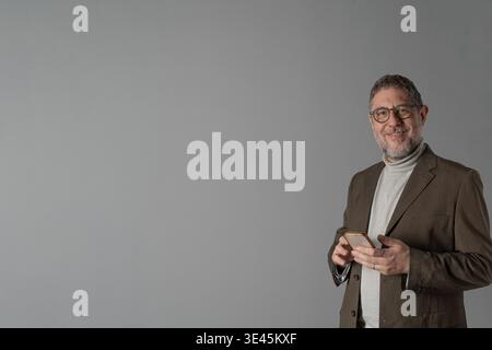 Ein lächelnder Mann mittleren Alters mit Brille und Bart hält ein Smartphone, während er vor einem grauen Hintergrund mit viel Kopierraum steht und Sen repräsentiert Stockfoto