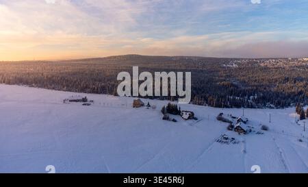 Schnee bedeckt das Dorf Jizerka im Isergebirge. Die Häuser sind umgeben von einer weiten weißen Fläche. Die Sonne geht auf und wirft Licht auf die verschneite Landschaft und Bäume. Stockfoto
