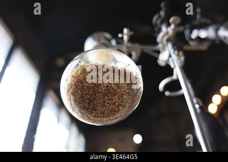 Nahaufnahme eines Glasbehälters gefüllt mit dunkel gerösteten Kaffeebohnen, montiert auf industriellen Metallgeräten in schwach beleuchteter Rösterei, die warm und invi ausdrücken Stockfoto