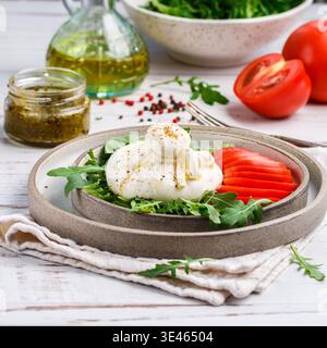 Cremiger italienischer Burrata-Käse, serviert mit frischer Rucola, Tomaten, Olivenöl, Basilikum-Pesto und frisch gemahlenem Pfeffer auf der grauen Platte auf einem weißen Wald Stockfoto