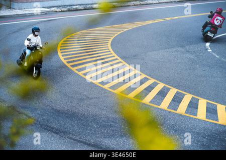 Hualien, Taiwan - 26. Februar 2023: Ein Roller und Lebensmittelkurier kreuzen auf einer geschwungenen gelben Straßenmarkierung, fotografiert von oben durch Laub. Stockfoto