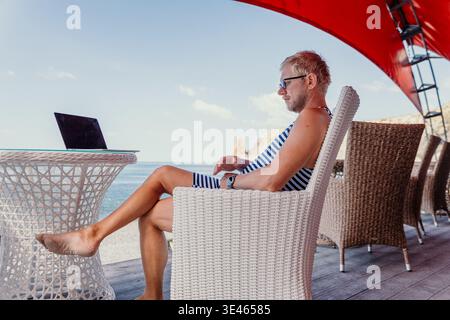 Digital Nomad Beach Freelancer arbeitet auf seinem Laptop auf einer sonnigen Terrasse am Meer während seiner Sommerferien ins Ausland Stockfoto