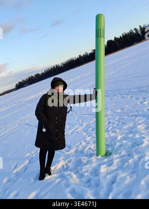 Eine zufriedene Frau, die in einem schwarzen Wintermantel, einem grünen Strickmütze und einem Schal gekleidet ist, posiert mit einem warmen Lächeln, während sie sich gegen eine große, hellgrüne Marke lehnt Stockfoto