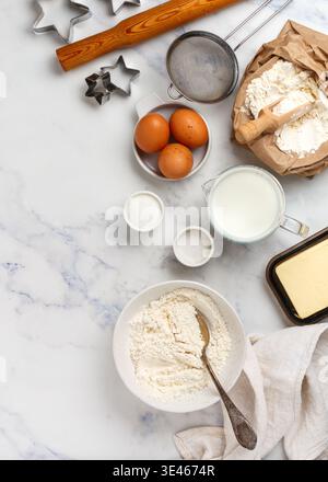 Teller mit Weizenmehl und Zutaten für die Zubereitung von Teig für hausgemachte Kekse oder Lebkuchen - Eier, Milch, Salz, Zucker, Butter auf Marmorhintergrund Stockfoto