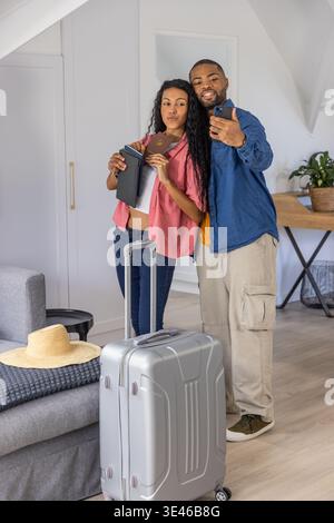 Ein Paar posiert für Selfie mit einem Mann, der ein Telefon hält, und einer Frau, die einen Pass in der Nähe eines Koffers auf dem Dachboden hält. Gepäck, Reisende, Verpackung, Loft, modern, fröhlich Stockfoto