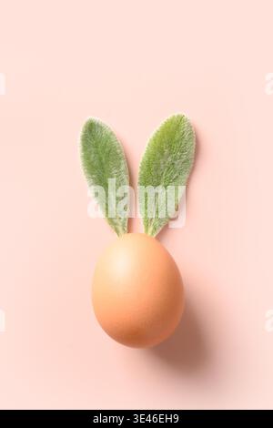 Osterbraunes Ei mit Blättern als Häschen auf rosa Hintergrund. Kreatives Konzept. Vertikale Grußkarte für Ostern. Blick von oben. Stockfoto