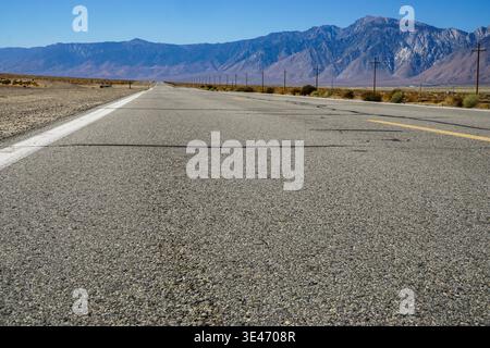 Flacher Blick auf die leere Wüstenautobahn, die in entfernte Berge mit Stromleitungen und trockener Landschaft unter klarem Himmel führt Stockfoto