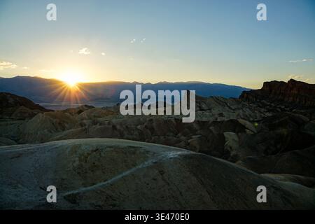 Sonnenuntergang über erodierten Wüstenbadlands und zerklüfteten Hügeln mit Bergketten in der Ferne unter klarem Himmel. Stockfoto