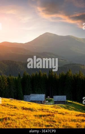 Bergtal mit Holzhütten umgeben von Wald und warmes Abendlicht. Ländliche Landschaft im Hochland mit friedlicher Golden Hour Stimmung Stockfoto