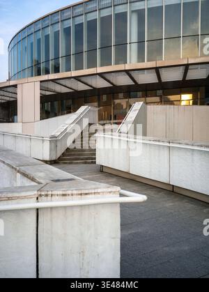 Modernes zylindrisches Glasgebäude mit Betontreppen und einem leeren Fußgängerweg. Die Szene repräsentiert zeitgenössische Unternehmensarchitektur, Stockfoto