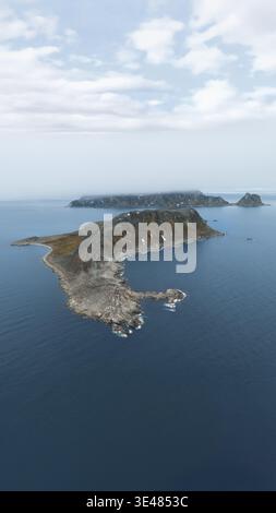Blick aus der Vogelperspektive auf eine zerklüftete Felseninsel mit steilen Klippen und Schneestücken, umgeben vom tiefblauen Wasser des Arktischen Ozeans Grönland. Stockfoto